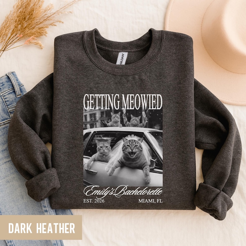 Pu&ograve; includere: Felpa grigio scuro con la scritta "GETTING MEOWIED" e un'immagine in bianco e nero di gatti in un'auto. La felpa include anche "Emily's Bachelorette" e "EST. 2026 MIAMI, FL".