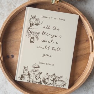 May include: A cream-coloured journal with the text "Letters to my Mom" and "all the things I wish I could tell you." The cover features illustrations of owls, a lantern, a mushroom, rabbits, and deer. The journal is on a wooden tray.