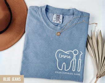 May include: A light blue t-shirt with a white graphic of a tooth and dental tools. The name "Emma" is inside the tooth graphic, and the text "YOUR CORPORATE NAME" is below. A gold necklace is draped across the shirt.
