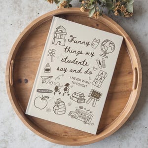Op de afbeelding: Een crèmekleurig notitieboek met harde kaft voorzien van donkerbruine lijntekeningen van schoolgerelateerde items en de tekst "Funny things my students say and do" gevolgd door "I NEVER WANT TO FORGET" met tekeningen van een bus en boeken.