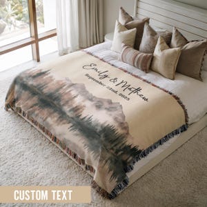 May include: A beige woven throw blanket with a mountain and forest watercolour design. The blanket has a fringed edge and features the names "Emily & Mathew" and the date "September 22nd, 2025".
