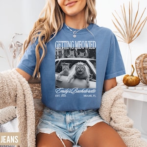 Pu&ograve; includere: T-shirt blu a maniche corte con la scritta "GETTING MEOWIED" sopra un'immagine in bianco e nero di gatti in un'auto. Il testo "Emily's Bachelorette" e "EST. 2026 MIAMI, FL" sono sotto. La modella indossa pantaloncini di jeans blu.