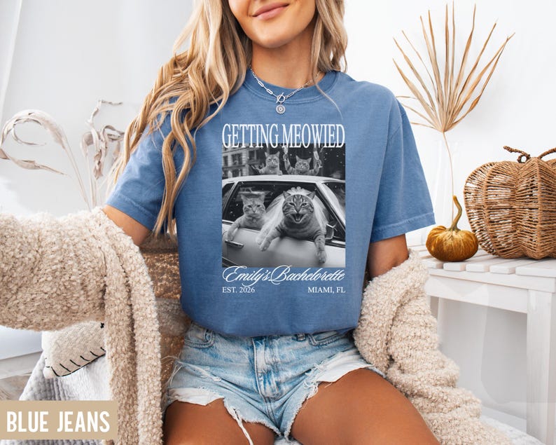 Pu&ograve; includere: T-shirt blu a maniche corte con la scritta "GETTING MEOWIED" sopra un'immagine in bianco e nero di gatti in un'auto. Il testo "Emily's Bachelorette" e "EST. 2026 MIAMI, FL" sono sotto. La modella indossa pantaloncini di jeans blu.