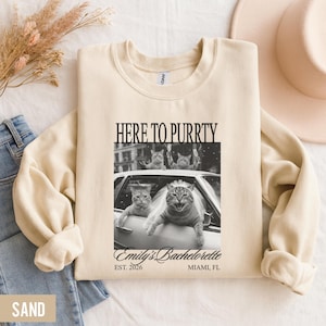 Pu&ograve; includere: Felpa color sabbia con la scritta "HERE TO PURRTY" sopra un'immagine in bianco e nero di gatti in un'auto. L'immagine include il testo "Emily's Bachelorette" e "EST. 2026 MIAMI, FL".