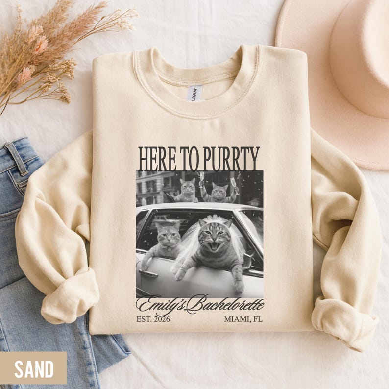 Pu&ograve; includere: Felpa color sabbia con la scritta "HERE TO PURRTY" sopra un'immagine in bianco e nero di gatti in un'auto. L'immagine include il testo "Emily's Bachelorette" e "EST. 2026 MIAMI, FL".