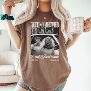 Pu&ograve; includere: T-shirt marrone con la scritta "GETTING MEOWIED" sopra un'immagine in bianco e nero di gatti in un'auto. L'immagine include anche "Emily's Bachelorette" e "EST. 2026 MIAMI, FL". La persona tiene in mano una tazza rosa.