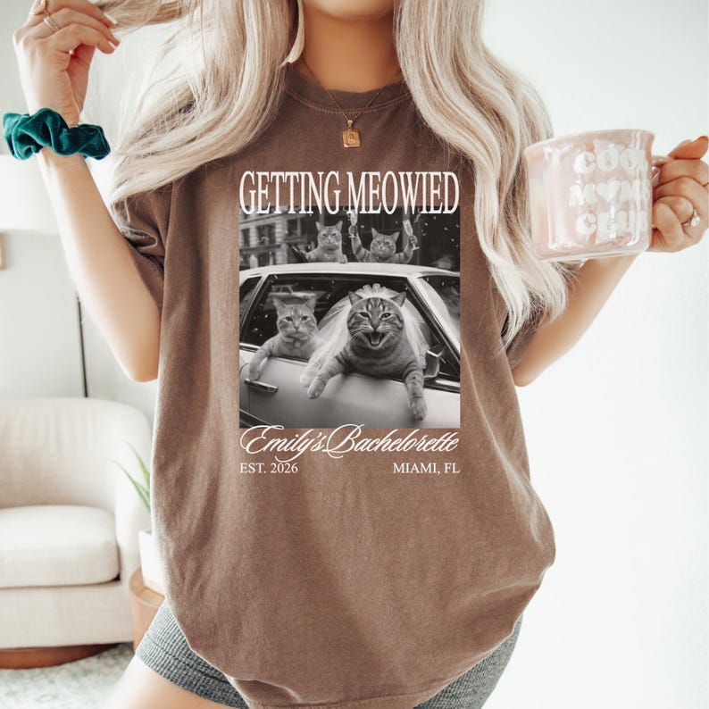 Pu&ograve; includere: T-shirt marrone con la scritta "GETTING MEOWIED" sopra un'immagine in bianco e nero di gatti in un'auto. L'immagine include anche "Emily's Bachelorette" e "EST. 2026 MIAMI, FL". La persona tiene in mano una tazza rosa.