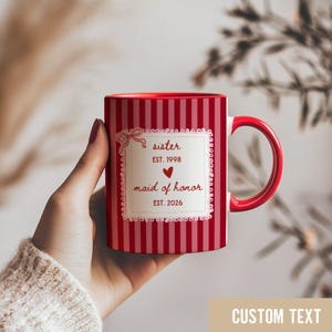 Puede incluir: Taza de cerámica a rayas rojas y blancas con asa e interior rojos. La taza presenta una etiqueta decorativa con las palabras "sister" y "maid of honor" y los años 1998 y 2026. El texto "CUSTOM TEXT" está en la parte inferior.