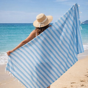 May include: A blue and white striped beach towel with fringed edges is held up on a sunny beach. The towel is rectangular and has vertical stripes. A straw hat with a ribbon is worn by a person standing on the beach.