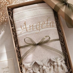 Op de afbeelding: Een beige en wit gestreepte deken met de naam "Angela" geborduurd in goudkleurig garen, gepresenteerd in een bruine geschenkdoos. De deken heeft een franjesrand en is vastgebonden met een groen lint. Een klein kaartje leest "Especially for you."