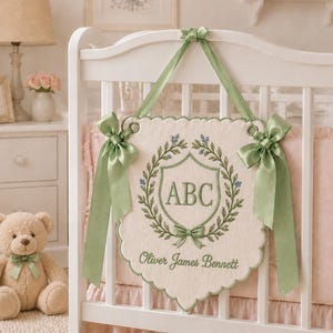 Op de afbeelding: Een crèmekleurige stoffen wandhanger met geschulpte randen, voorzien van een geborduurde groene lauwerkrans rond de letters ABC en de naam Oliver James Bennett. De banner hangt aan saliegroene satijnen linten die in strikken zijn geknoopt.