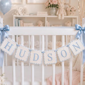 以下が含まれることがあります： スカラップエッジの布製スクエアに青い刺繍で「HUDSON」と綴られた白いベビーベッド用バナー。バナーは白いベビーベッドに取り付けられており、上部レールの両端には水色のサテンリボンが蝶結びで結ばれています。