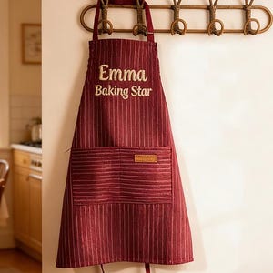 Puede incluir: Un delantal burdeos con rayas verticales y un bolsillo delantero cuelga de un perchero decorativo. El delantal tiene el texto "Emma Baking Star" bordado. El perchero es de metal dorado.