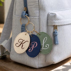 Peut inclure: Un sac à dos à rayures bleu clair et blanc avec trois porte-clés en tissu de forme ovale attachés à la tirette de la fermeture éclair. Les porte-clés sont beige avec un K marron, bleu marine avec un P rose et vert sauge avec un L blanc.