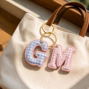 May include: A cream-colored canvas tote bag with brown leather handles features two gold-toned metal rings attached to the handle. Hanging from the rings are two padded fabric letter charms, one blue and white gingham letter G and one pink and white gingham letter M.