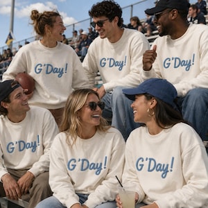 May include: A cream-colored crewneck sweatshirt featuring the text G'Day! embroidered in blue script lettering across the chest. The garment has long sleeves, a ribbed neckline, and a relaxed fit, constructed from a soft, knit fabric material.