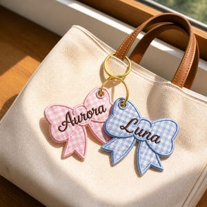 May include: A cream-colored canvas tote bag with brown leather handles features two embroidered gingham bow keychains attached to a gold ring. One bow is pink with the name Aurora, and the other is light blue with the name Luna in script.