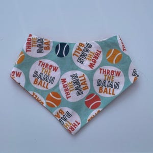 Puede incluir: Un pañuelo turquesa para perros con un patrón repetido de círculos blancos, cada uno con un gráfico de pelota de tenis y el texto "THROW THE DAMN BALL" en naranja y blanco. También hay un gráfico de pelota de tenis.