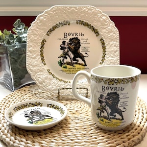 May include: A white ceramic set comprising a decorative plate, a small saucer, and a mug, each featuring a vintage Bovril advertisement. The design depicts a person wrestling a lion with the text Bovril for colds, chills and influenza.