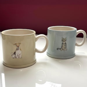 May include: Two ceramic mugs. One is cream-coloured with a dog illustration, the other is light blue with a cat illustration. Both mugs have white handles and are on a white surface.