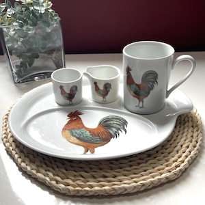 Peut inclure: Un service à petit-déjeuner en céramique blanche avec un motif de coq. L'ensemble comprend un grand plat ovale, une tasse, une crémière et deux petites tasses. Le coq est représenté en marron, rouge, bleu et vert. L'ensemble est posé sur un set de table tressé.