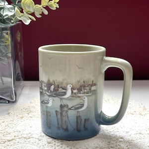 May include: A ceramic mug with a coastal design. The mug features a scene with seagulls perched on wooden posts, a harbor town, and a gradient of blue and beige hues. The mug has a large, square handle.