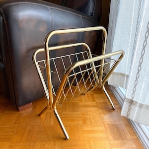 May include: A polished gold-tone metal magazine rack featuring a structured frame with a wire grid design. The stand is constructed with two arched side supports and a central V-shaped basket for holding reading materials in an upright position.
