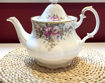 Pode incluir: Um bule de porcelana branca com um delicado padrão floral de rosas cor-de-rosa, roxas e verdes. O bule tem uma borda recortada, uma alça curva, um remate de tampa pontiagudo e detalhes em ouro ao longo das bordas e do bico.