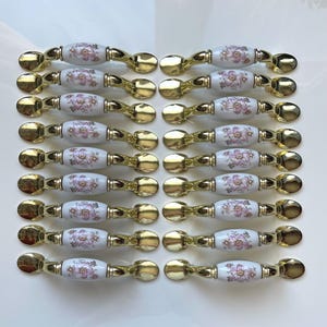 May include: A collection of cabinet pulls with a gold finish and white ceramic centers. The ceramic sections are decorated with pink floral patterns. The pulls are arranged in neat rows, showcasing their design and quantity.