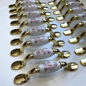 May include: Cabinet handles with a gold finish and white ceramic centres. The ceramic has a floral design in pink and brown. The handles are arranged in rows, highlighting their decorative style and potential for home decor.