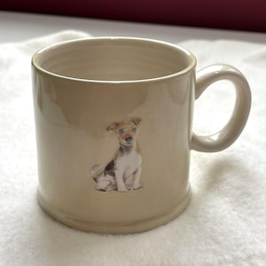 Puede incluir: Una taza de cerámica beige con un asa redondeada. La taza presenta una ilustración de un perro sentado en marrón, blanco y beige. La taza tiene un acabado brillante y es adecuada para bebidas frías o calientes. La taza mide unos 10 cm de alto.