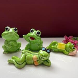Può includere: Quattro statuette di rana in ceramica verde in varie pose. Una rana è seduta, un'altra saluta e due sono sdraiate. Una rana sdraiata indossa occhiali da sole e un bikini. Le rane sono su una superficie bianca con uno sfondo bordeaux.