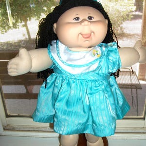 May include: A doll with a soft fabric body, light skin tone, and dark yarn hair styled with a blue ribbon. The doll wears a bright turquoise dress featuring a white collar with a small embroidered detail and a subtle vertical striped pattern.
