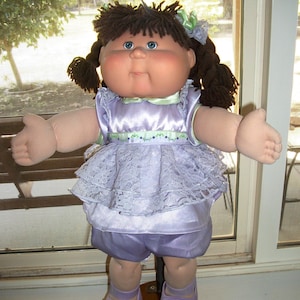 May include: A soft-sculpture doll with dark brown yarn hair styled in pigtails with light green bows. The doll has blue eyes, a round face, and wears a lavender satin dress with a lace overlay, a light green ribbon trim, and matching lavender shoes.