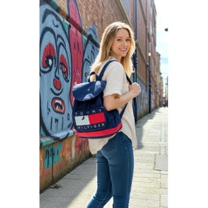 Puede incluir: Una mochila azul marino con base roja y logotipo blanco, rojo y azul. La mochila tiene cierre de solapa y correas ajustables. El texto "TOMMY HILFIGER" está impreso en la parte delantera. Una persona lleva la mochila.