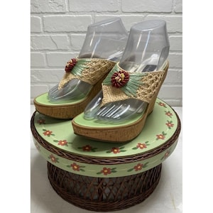 May include: Pair of wedge sandals with woven raffia straps and a floral accent. The sandals have a green footbed and a cork wedge heel. The sandals are displayed on a green and brown floral pedestal.