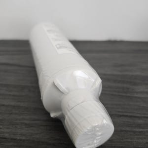 Könnte beinhalten: Eine weiße zylindrische Flasche, in klarem Kunststoff versiegelt, liegt auf einer dunkelgrauen Holzoberfläche. Die Flasche hat einen abgerundeten Boden und einen weißen Verschluss. Die transparente Verpackung ermöglicht einen Blick auf das Produkt.