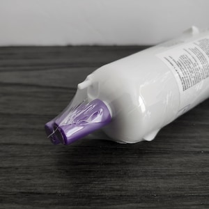 Puede incluir: Un cartucho de filtro de agua cilíndrico blanco, sellado en plástico transparente. Los extremos son morados. El filtro está sobre una superficie de madera gris oscuro.