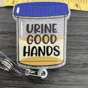May include: A glittery badge reel with a clear container design, featuring the words "URINE GOOD HANDS" in black lettering. The container has a blue lid and a yellow liquid inside. Includes a retractable cord and a metal clip.