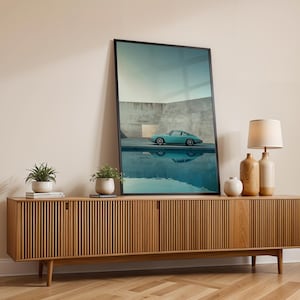 May include: A framed art print featuring a teal sports car reflected in water, displayed on a wooden cabinet. The cabinet has a slatted design and is flanked by decorative vases, a lamp, and potted plants. The room has a neutral color palette.