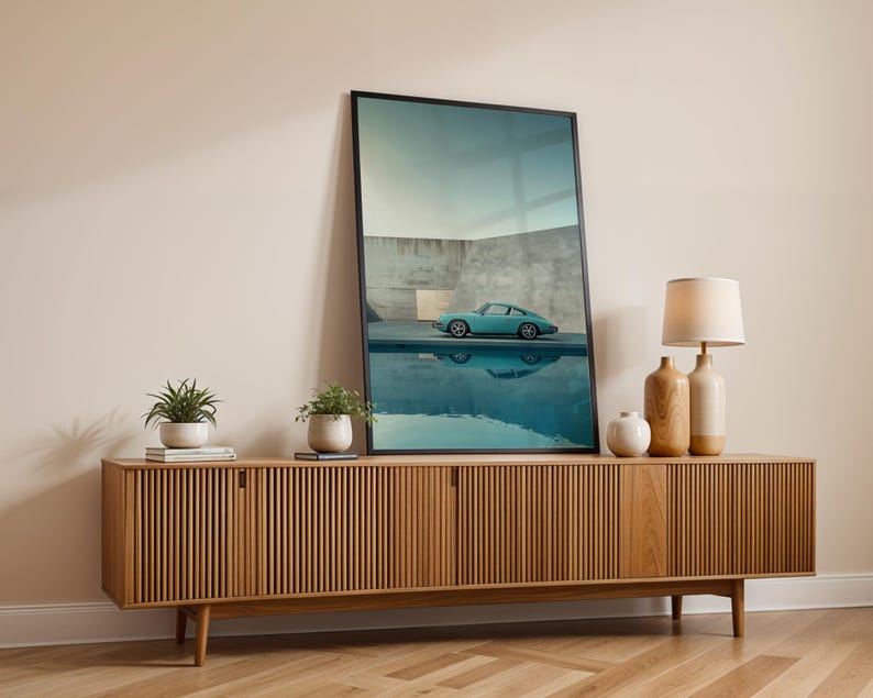May include: A framed art print featuring a teal sports car reflected in water, displayed on a wooden cabinet. The cabinet has a slatted design and is flanked by decorative vases, a lamp, and potted plants. The room has a neutral color palette.