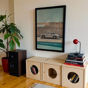May include: A living room scene featuring a framed car print, a red desk lamp, and three wooden storage cubes with circular openings. A potted plant and a black speaker are also visible.