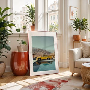May include: A framed art print featuring a yellow sports car parked by a pool with a mountain backdrop. The print is displayed in a bright, airy living room with potted plants, a red side table, and a cream-colored sofa.