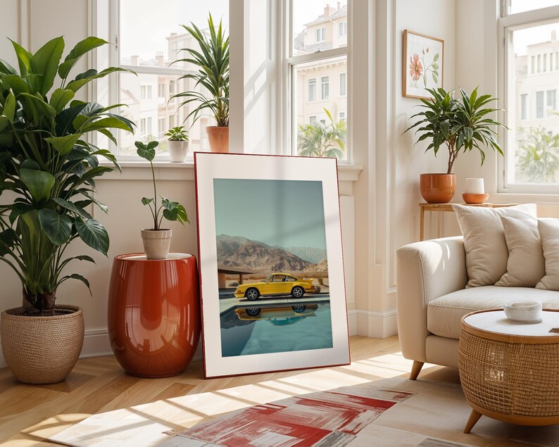 May include: A framed art print featuring a yellow sports car parked by a pool with a mountain backdrop. The print is displayed in a bright, airy living room with potted plants, a red side table, and a cream-colored sofa.