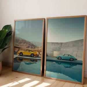 May include: Two framed art prints featuring vintage sports cars. One print shows a yellow car near mountains, the other a teal car near a pool. Both prints have wooden frames and are displayed on a wooden floor.