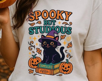 May include: A white short-sleeved t-shirt featuring a graphic print of a black cat wearing a purple witch hat, sitting on a stack of books labeled Spells, surrounded by orange pumpkins and the text Spooky But Studious in orange and teal.
