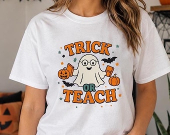 May include: A white short-sleeved t-shirt featuring a central graphic of a ghost wearing glasses, holding two books, and flanked by two orange pumpkins. The text reads "TRICK OR TEACH" in orange letters, surrounded by small stars and black bats.