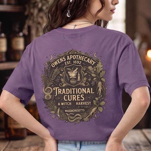 May include: A purple short-sleeve t-shirt featuring a graphic on the back with the text Owens Apothecary Est. 1692 Traditional Cures A Witch Harvest Massachusetts, surrounded by illustrations of plants, a mortar and pestle, and a snake.