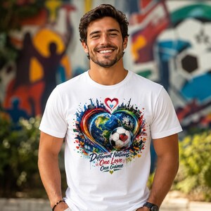 May include: A white short-sleeved t-shirt featuring a colorful graphic print of a heart-shaped earth, a soccer ball, and various global landmarks. The text "Different Nations One Love One Game" is printed in blue and red script below the graphic.