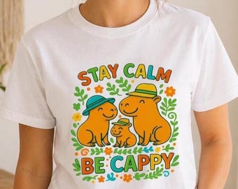 Puede incluir: Camiseta blanca con un gráfico colorido de tres capibaras de dibujos animados con sombreros, rodeados de hojas y flores. El texto "STAY CALM BE CAPPY" se muestra arriba y abajo de los capibaras.